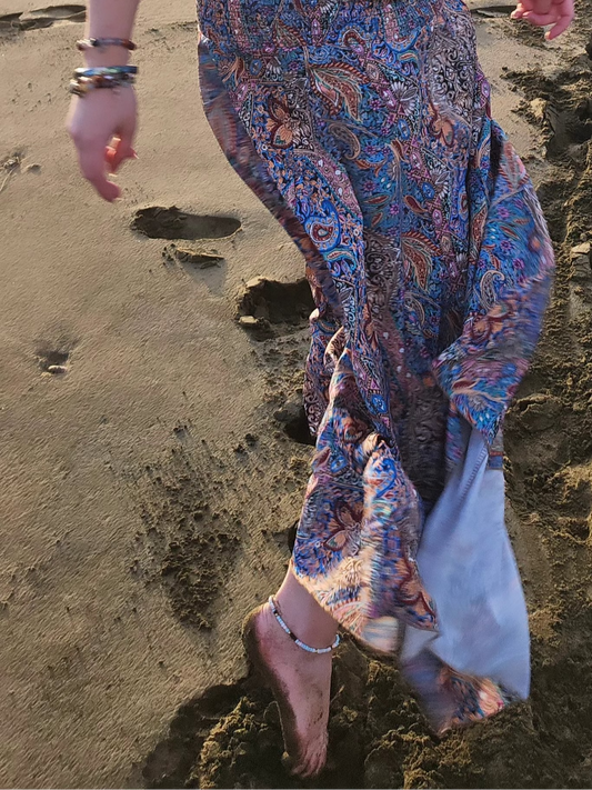 Person wearing colorful patterned skirt and anklet walking on a sandy beach.